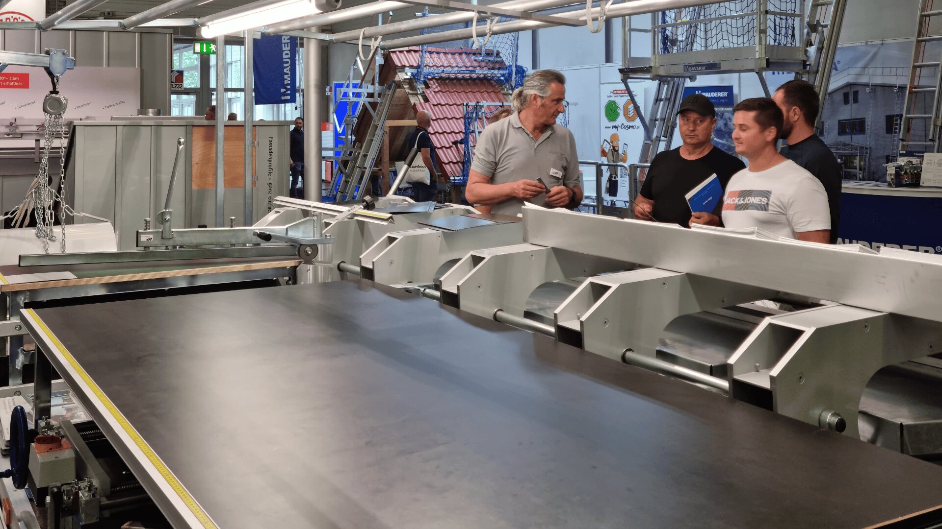 Vier Männer stehen und unterhalten sich neben einem großen industriellen Förderband oder einer Produktionsmaschine in einer Fabrik. Im Hintergrund sind Werkzeuge, Geräte und Schilder zu sehen.