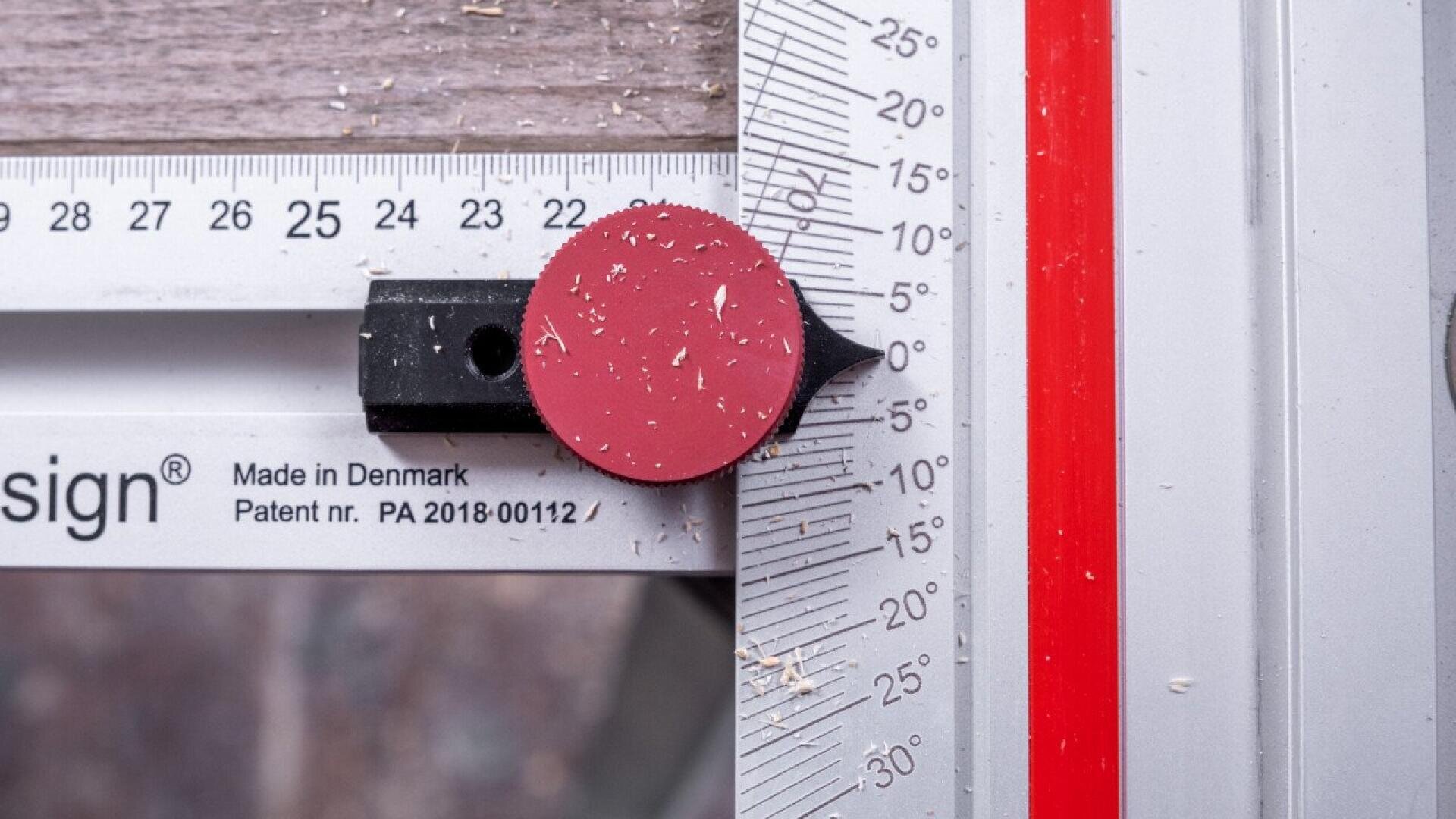 Eine Nahaufnahme eines Messwerkzeugs mit einer roten kreisförmigen Markierung, die in einem 45-Grad-Winkel auf einem metallischen Winkelmesser angebracht ist, wobei Holzspäne auf dem Werkzeug verstreut sind.