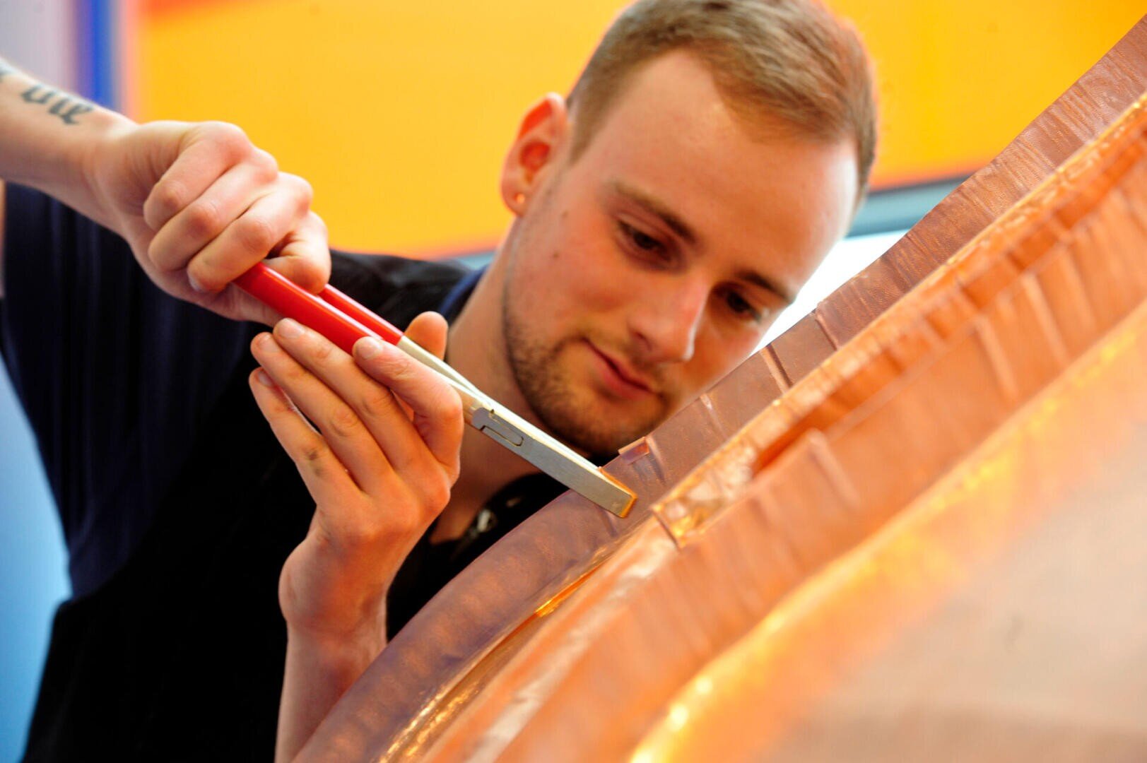 A man uses pliers to carefully shape or work on a large, curved metal surface, focusing intently on the task. The setting appears to be a workshop or industrial environment.