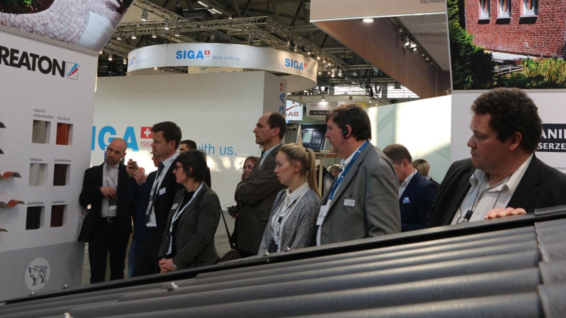 A group of people in business attire attentively listen to a man presenting roofing material samples at a trade show booth, with exhibition stands and banners visible in the background.