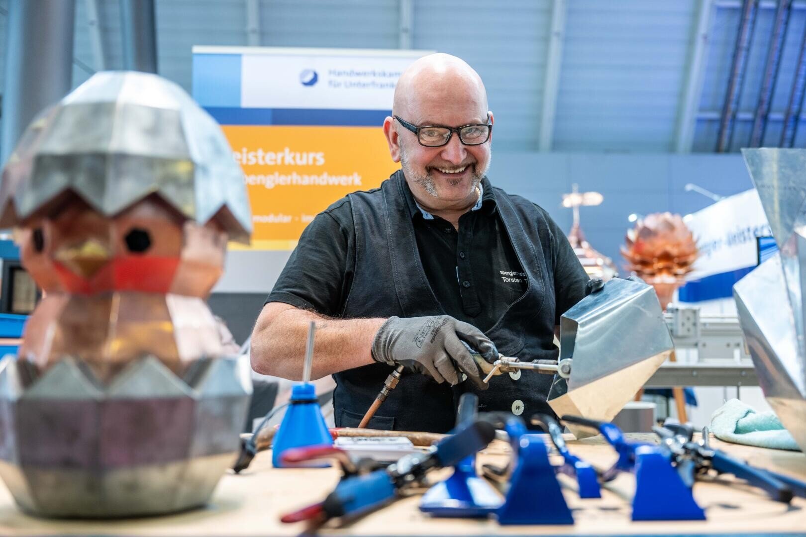 Ein lächelnder Mann mit Brille und Handschuhen arbeitet mit Metallwerkzeugen an einer Werkbank, um eine Skulptur zu schaffen. Im Vordergrund ist ein Metallküken zu sehen, das aus einem Ei schlüpft. Im Hintergrund sind Werkstattschilder und Geräte zu sehen.