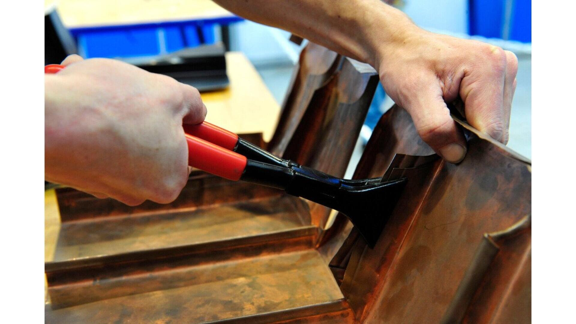 A person uses a pair of red-handled pliers to bend or shape a piece of sheet metal on a workbench.