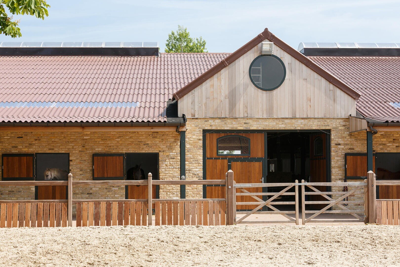 Ein moderner Stall aus Ziegeln und Holz mit einem braunen Ziegeldach, Holztoren und mehreren Boxen. Aus den Fenstern auf der linken Seite sind zwei Pferde zu sehen. Der Stallhof ist sandig und sonnenbeschienen.