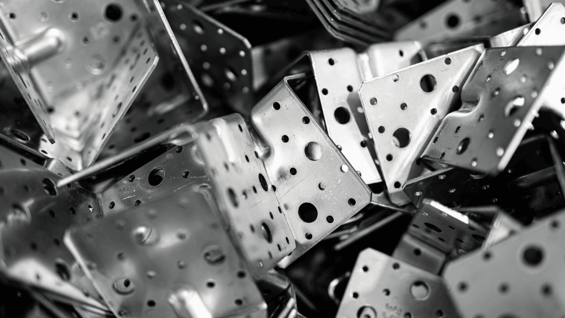 A close-up of a pile of shiny metal brackets with various holes and cutouts, arranged haphazardly. The image is in black and white, emphasizing the metallic texture and geometric shapes.