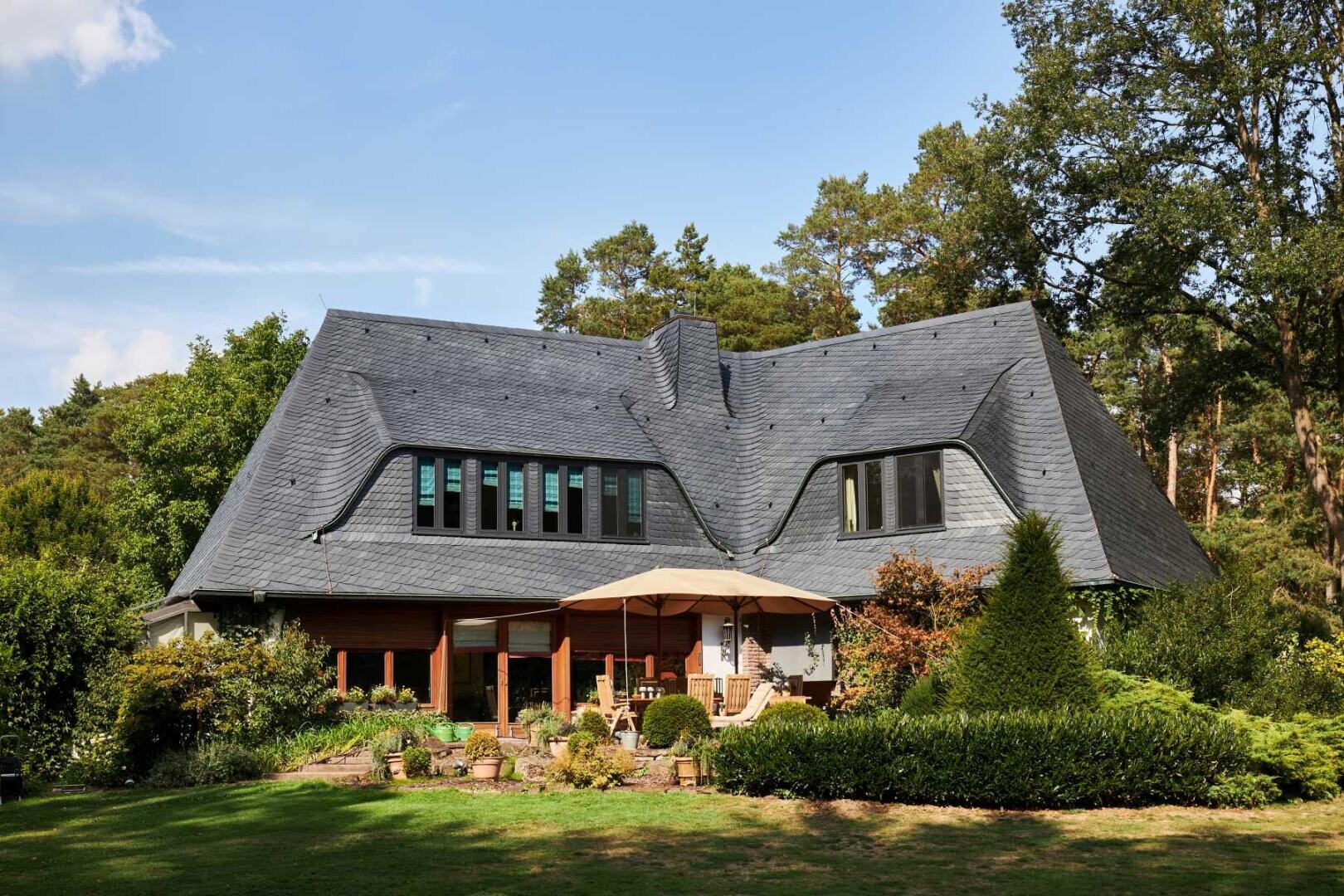 Ein großes Haus mit einem einzigartigen, gewellten dunklen Schieferdach, umgeben von üppigem Grün und Bäumen. Der Garten verfügt über Terrassenmöbel, einen Sonnenschirm und eine gepflegte Gartenanlage.