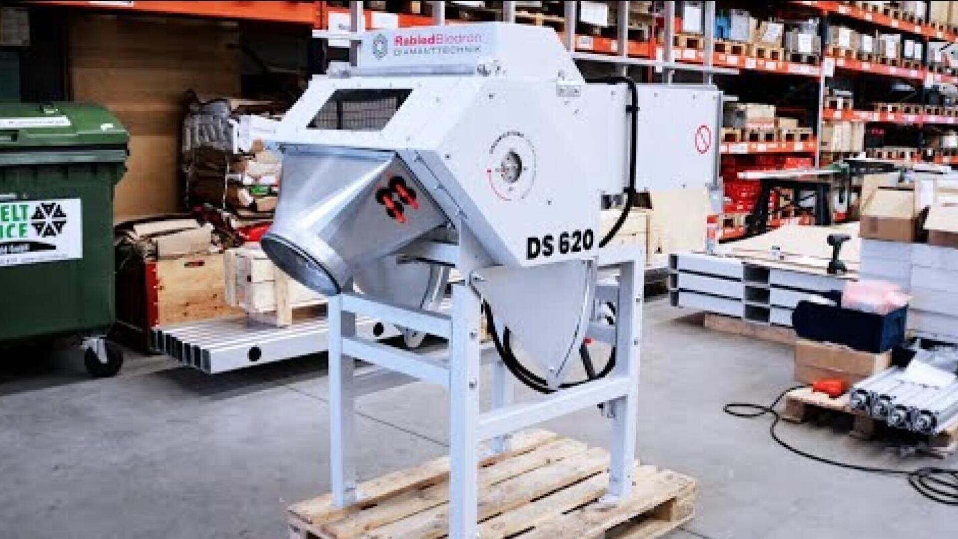 Industrial machine with the designation DS 620 and funnel-shaped outlet, mounted on a wooden pallet in a warehouse, surrounded by shelves with various crates and equipment.
