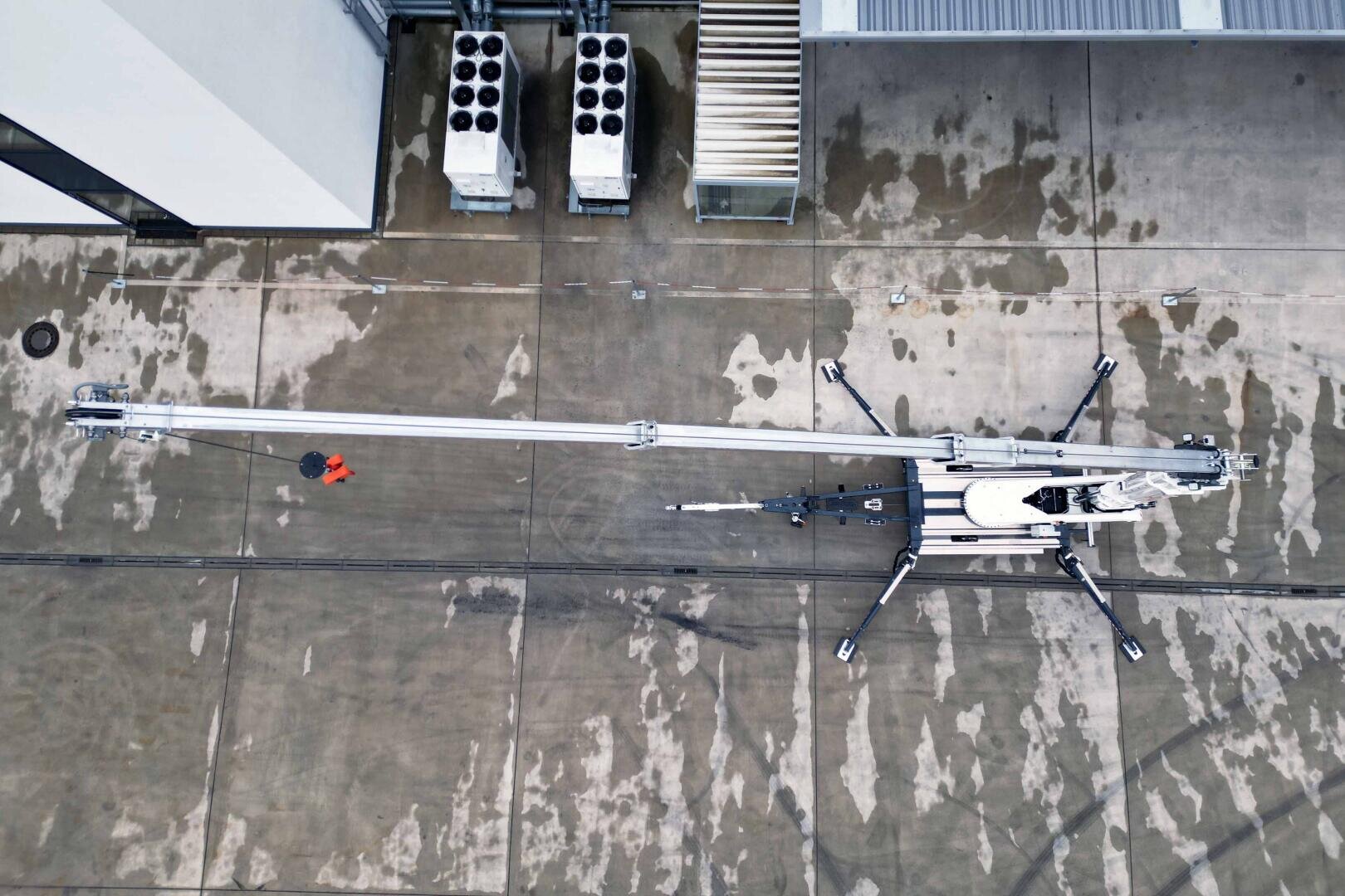 Aerial view of a person in an orange jacket standing on concrete near a large, extended crane or hoist with long stabilizing legs and metal equipment on the roof of a building.