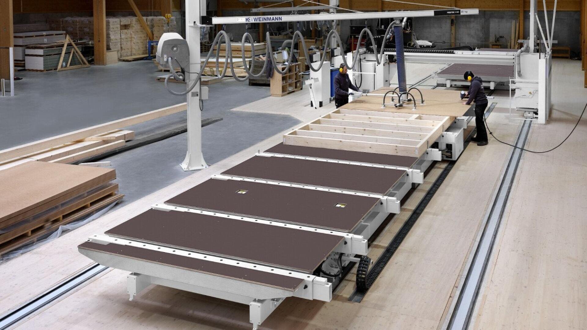 A large factory workshop with two workers operating machinery to assemble prefabricated wooden panels or modular structures, surrounded by wood panels, beams, and equipment.