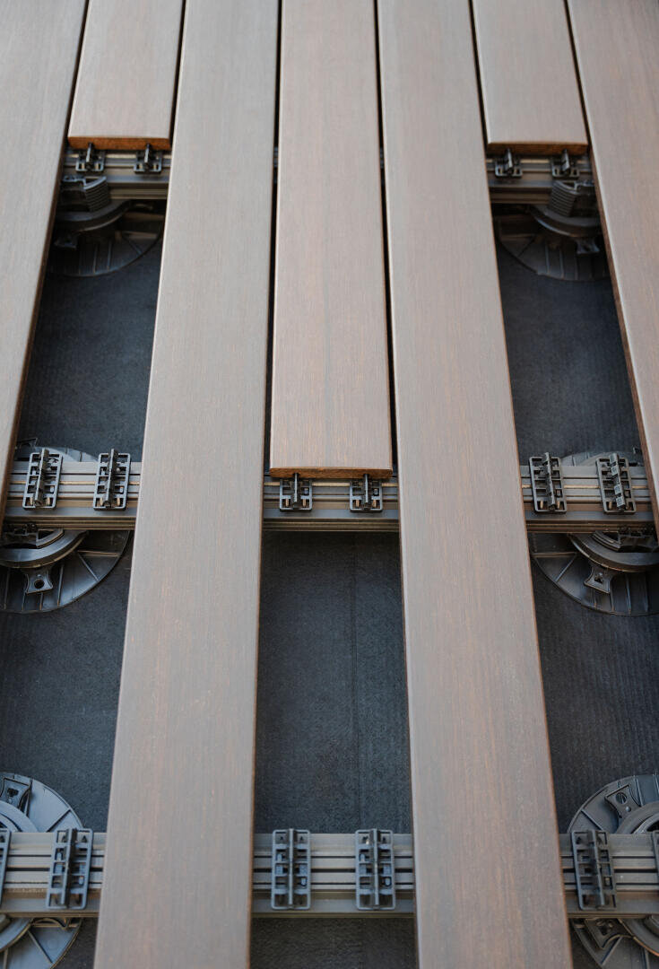 Nahaufnahme von braunen Terrassendielen aus Verbundwerkstoff, die teilweise auf verstellbaren schwarzen Kunststoffsockeln und Stützklammern verlegt sind, wodurch die darunter liegende Struktur und die Abstände zwischen den Dielen sichtbar werden.