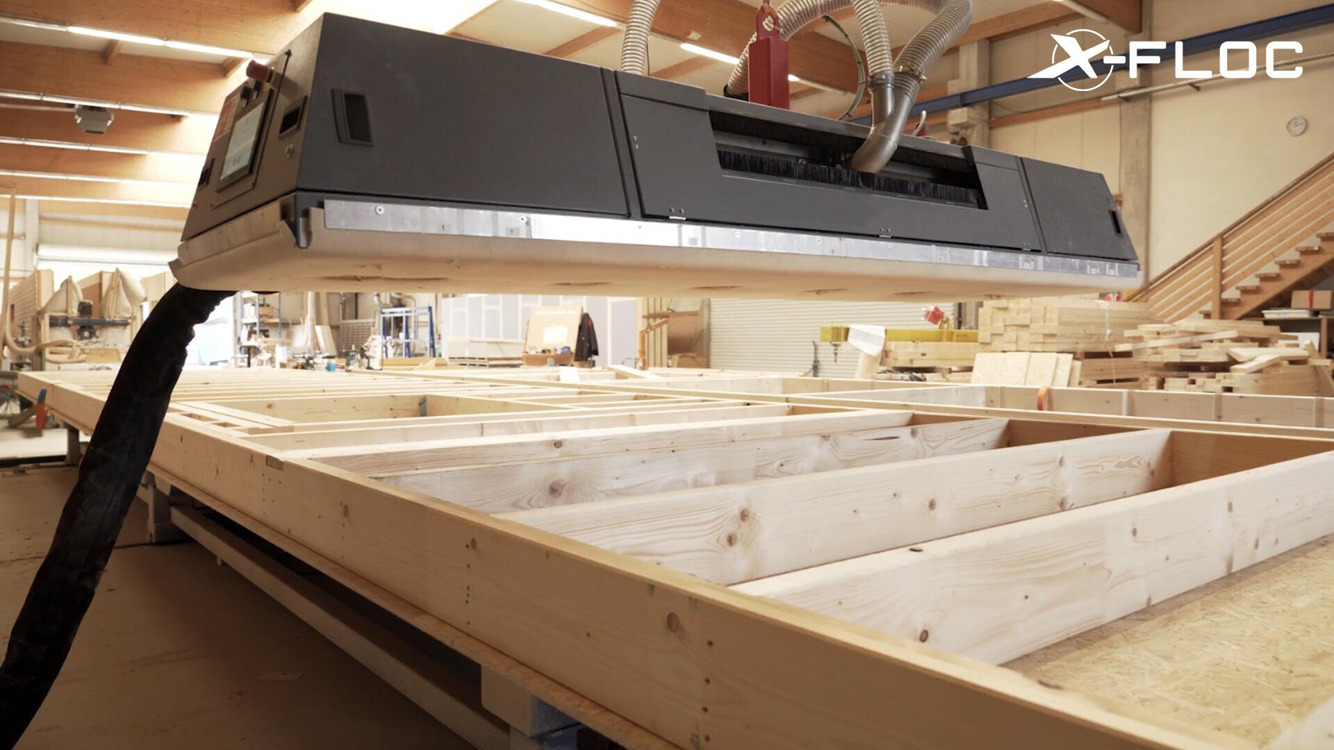 A large X-FLOC machine works on a wooden frame in a spacious, well-lit carpentry workshop filled with wood and equipment.