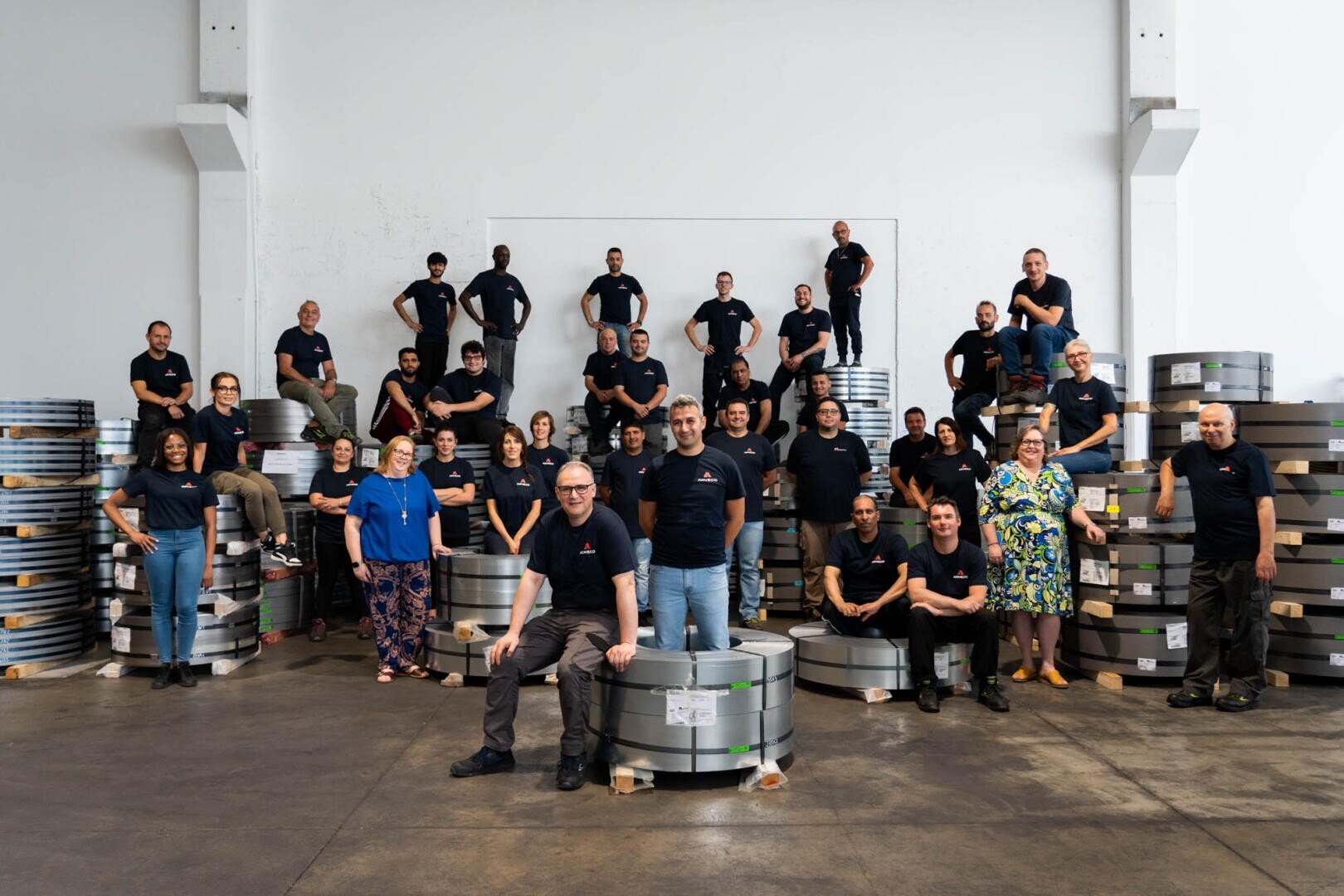 Eine Gruppe von etwa 35 Personen in passenden dunklen Hemden posiert lächelnd in einer Lagerhalle mit Stapeln von Metallspulen um sie herum. Einige stehen, andere sitzen oder knien vor dem Hintergrund einer weißen Wand.