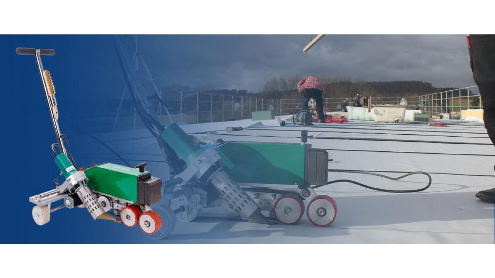 A green and black automated welding machine on a flat rooftop, with workers in the background installing roofing material under a cloudy sky.