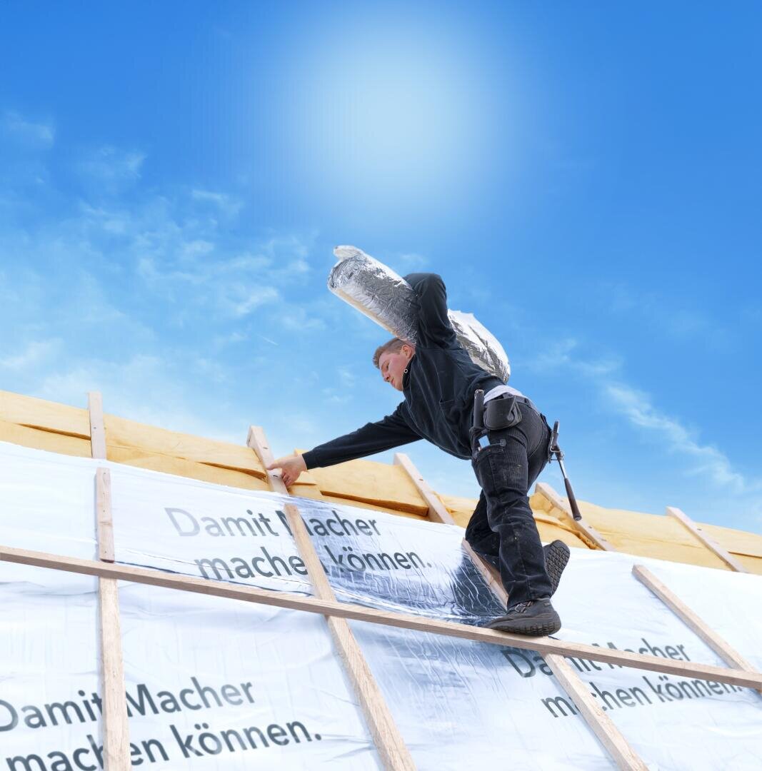 Ein Bauarbeiter in Sicherheitsausrüstung klettert unter blauem Himmel auf einen hölzernen Dachstuhl und trägt eine schwere Materialrolle auf seinen Schultern. Auf dem teilweise abgedeckten Dach steht ein deutscher Text.