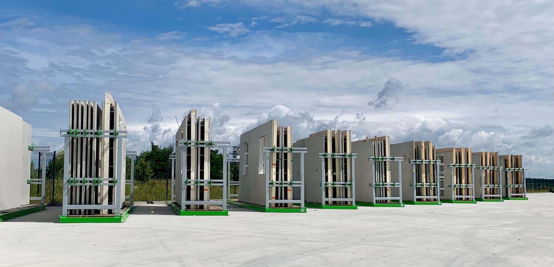 Vorgefertigte Betonwandplatten sind vertikal in grünen Metallgestellen auf einer großen Betonfläche im Freien gestapelt, mit einem bewölkten blauen Himmel und Bäumen im Hintergrund.