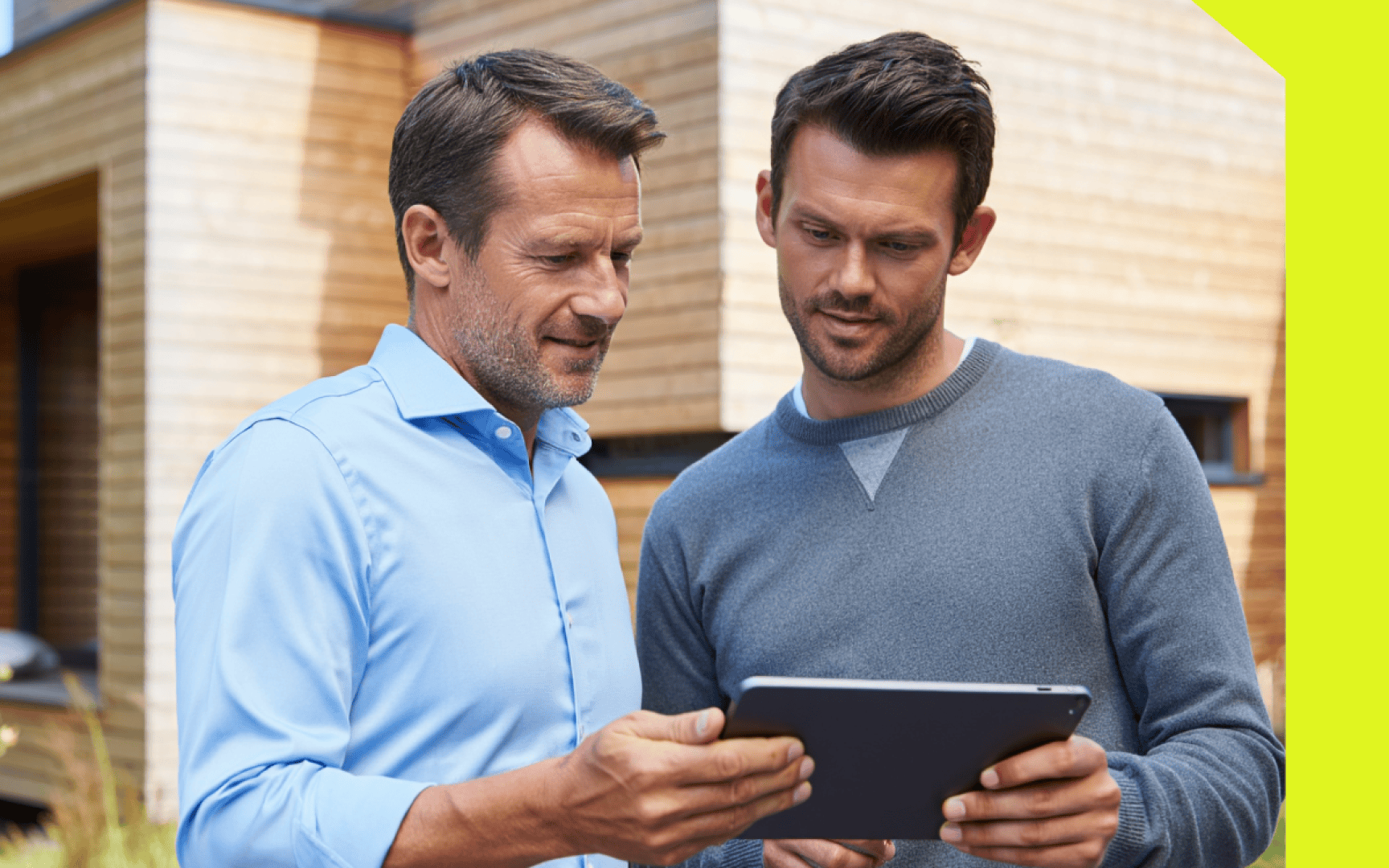Two men are standing outside a modern wooden building. Both are looking at a tablet that one of them is holding and seem to be discussing something on the screen.