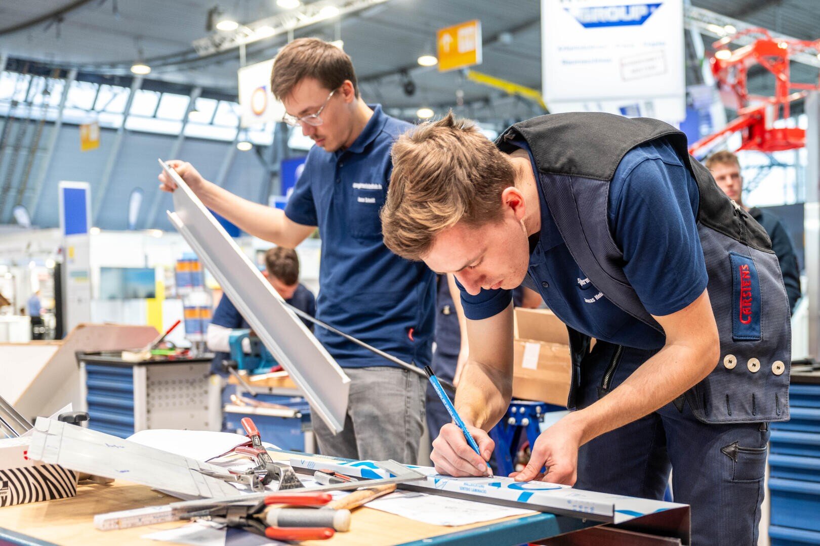 Zwei junge Männer in blauen Arbeitsuniformen montieren und prüfen elektrische Geräte an einer Werkbank in einer hellen, modernen Werkstatt. Einer schreibt auf ein Blatt, während der andere eine große Tafel hält. Werkzeuge und Drähte liegen verstreut auf dem Tisch.