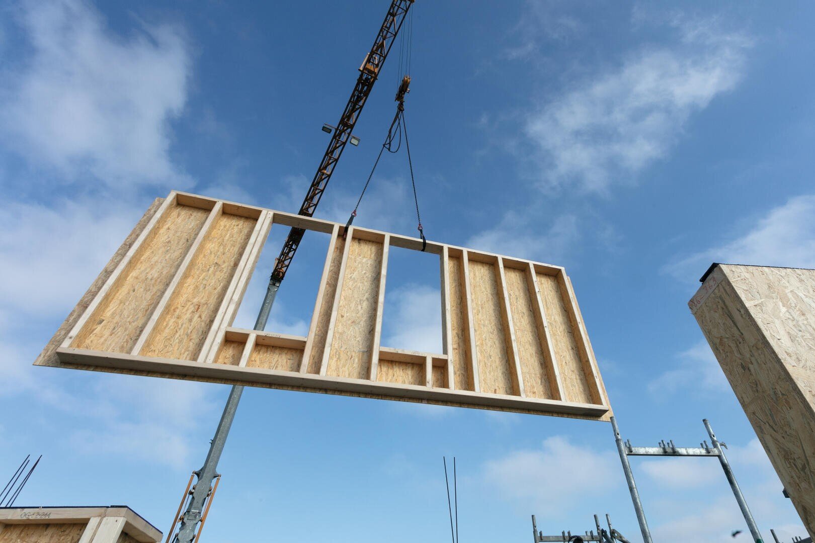 Ein Baukran hebt einen hölzernen Wandrahmen auf einer Baustelle unter blauem Himmel mit vereinzelten Wolken an seinen Platz.