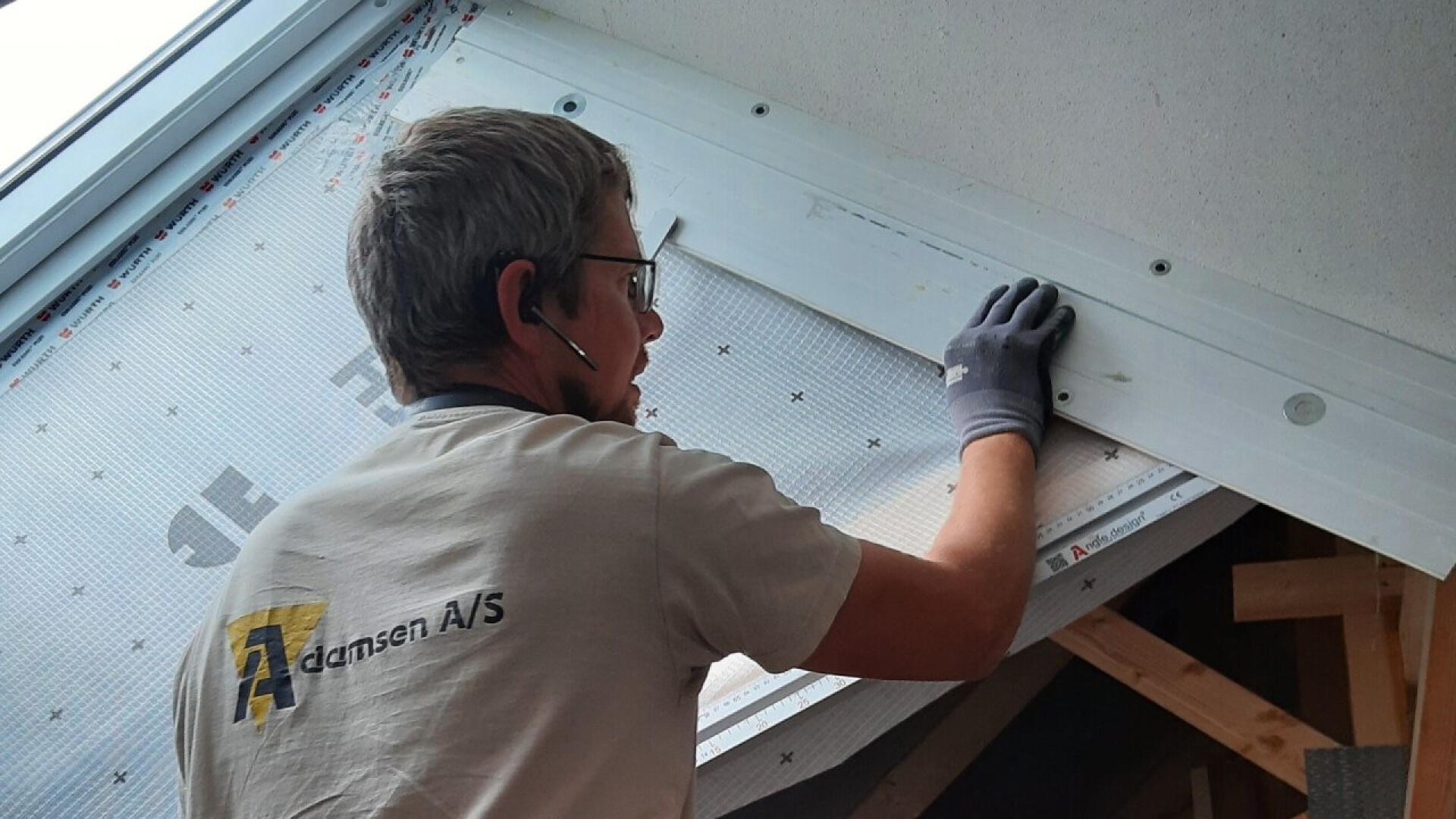 Eine Person mit Brille, Headset und Handschuh montiert eine Dämmung oder ein Paneel an der Dachschräge eines im Bau befindlichen Raumes. Auf dem T-Shirt steht Adnsen A/S. Licht fällt durch ein nahegelegenes Dachfenster ein.