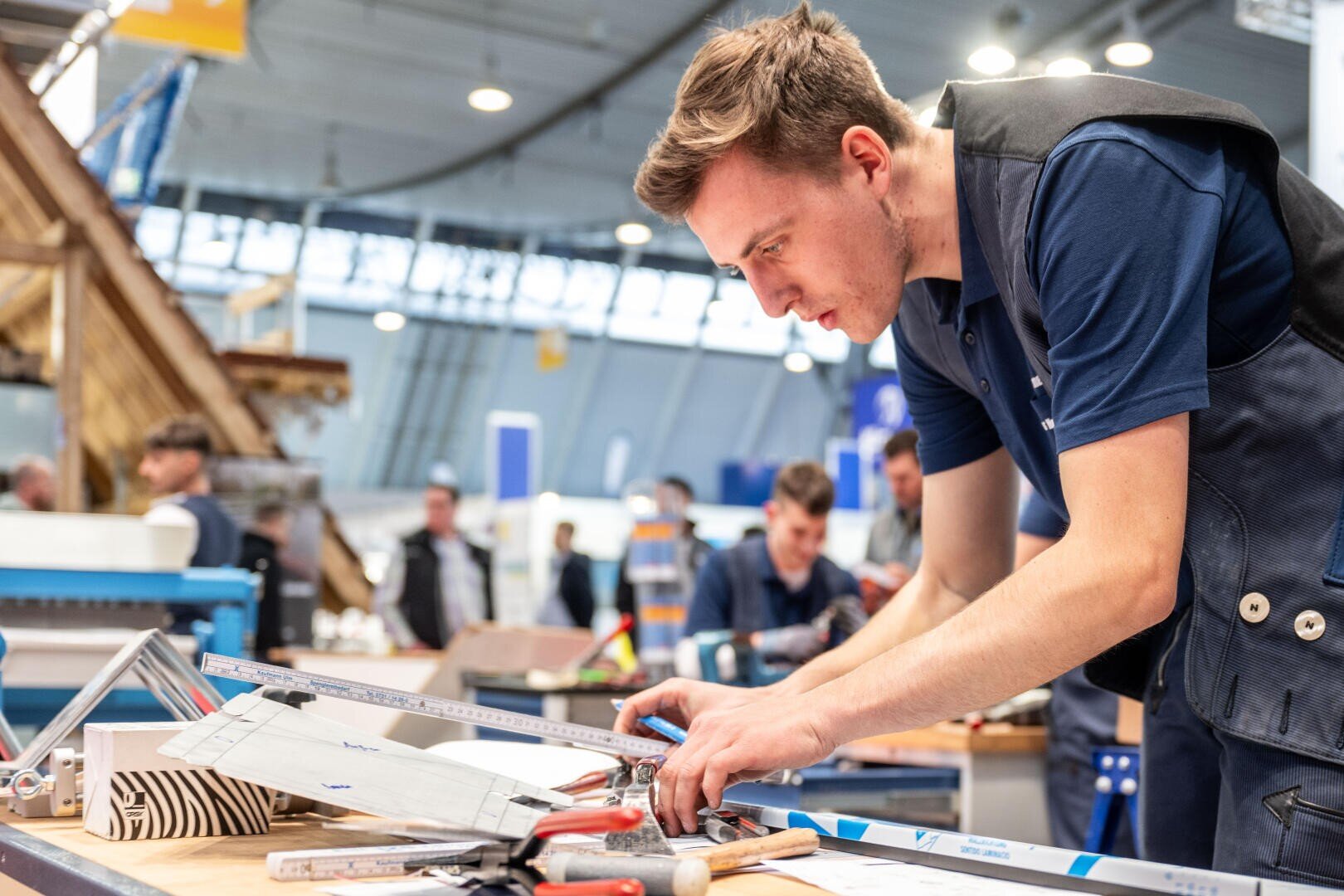 Ein junger Mann misst in einer Werkstatt ein Stück Material mit einem Lineal und einem Bleistift aus und konzentriert sich dabei sehr. Werkzeuge und Baumaterialien liegen verstreut auf dem Tisch. Im Hintergrund arbeiten andere Personen. Die Umgebung scheint ein Innenraum zu sein.