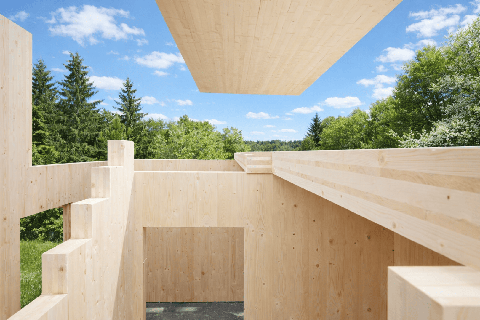 A modern wooden building under construction with exposed walls and beams outside, surrounded by green trees and a blue sky with scattered clouds. A large wooden plank is placed above the structure.
