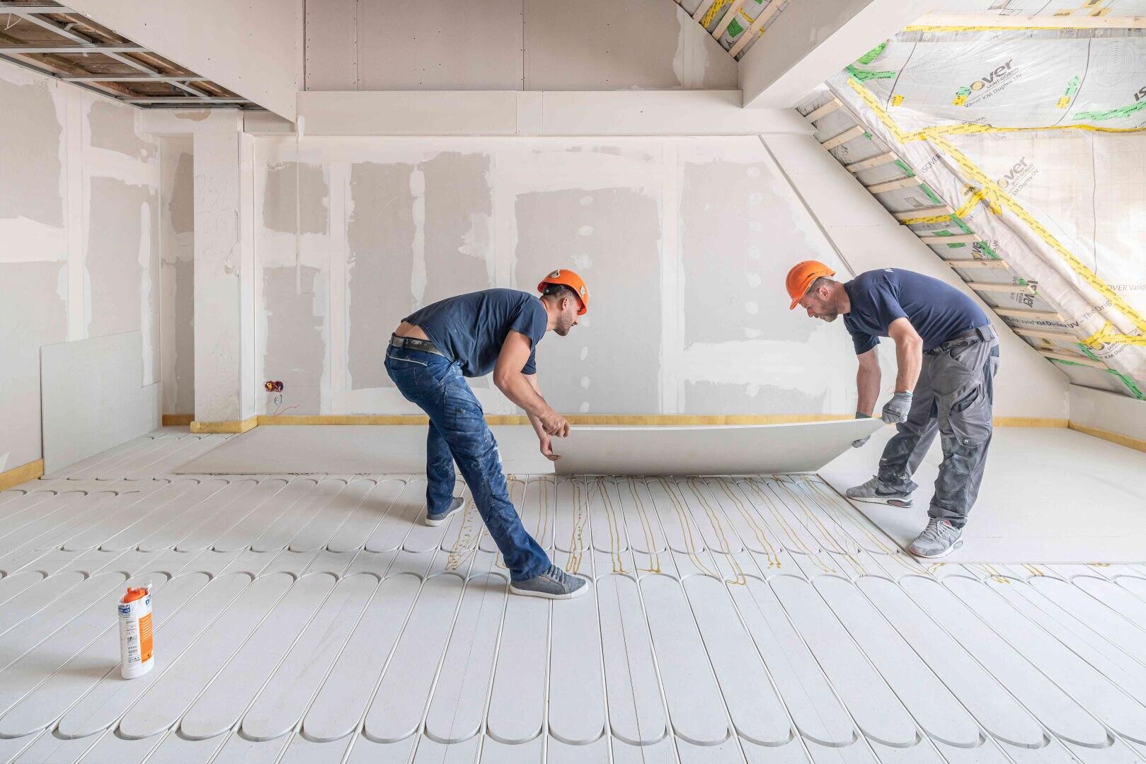 Two construction workers in orange helmets are installing a large drywall panel in a bright, unfinished room. The pipes for the underfloor heating are visible on the floor. The walls and ceiling are partially finished.