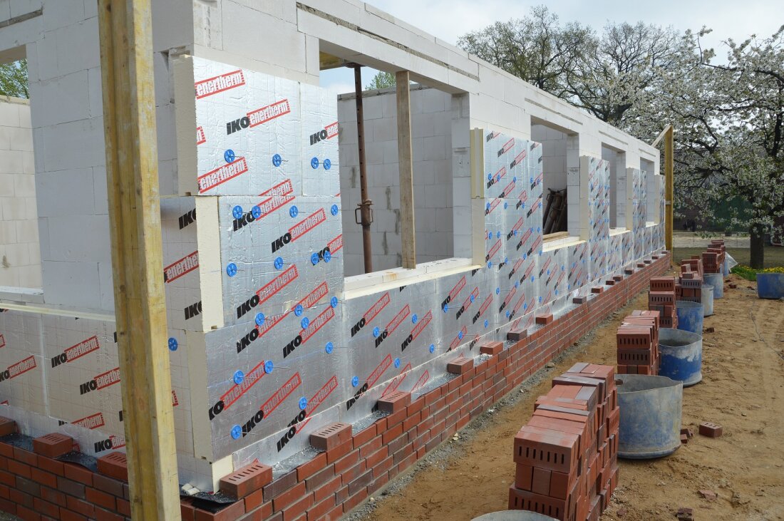 Ein teilweise errichtetes Gebäude, an dessen Außenwand Dämmplatten und mit Kunststoff ummantelte Ziegel angebracht werden. Mehrere unfertige Fensteröffnungen und Baumaterialien sind auf dem Boden zu sehen.