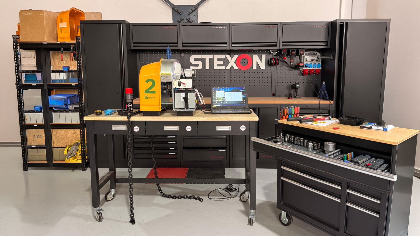 A well-organized workshop with black cabinets, a pegboard labeled STEXON, various tools, a yellow machine labeled 2, a laptop, and tool drawers. Shelves with boxes and bins are on the left.
