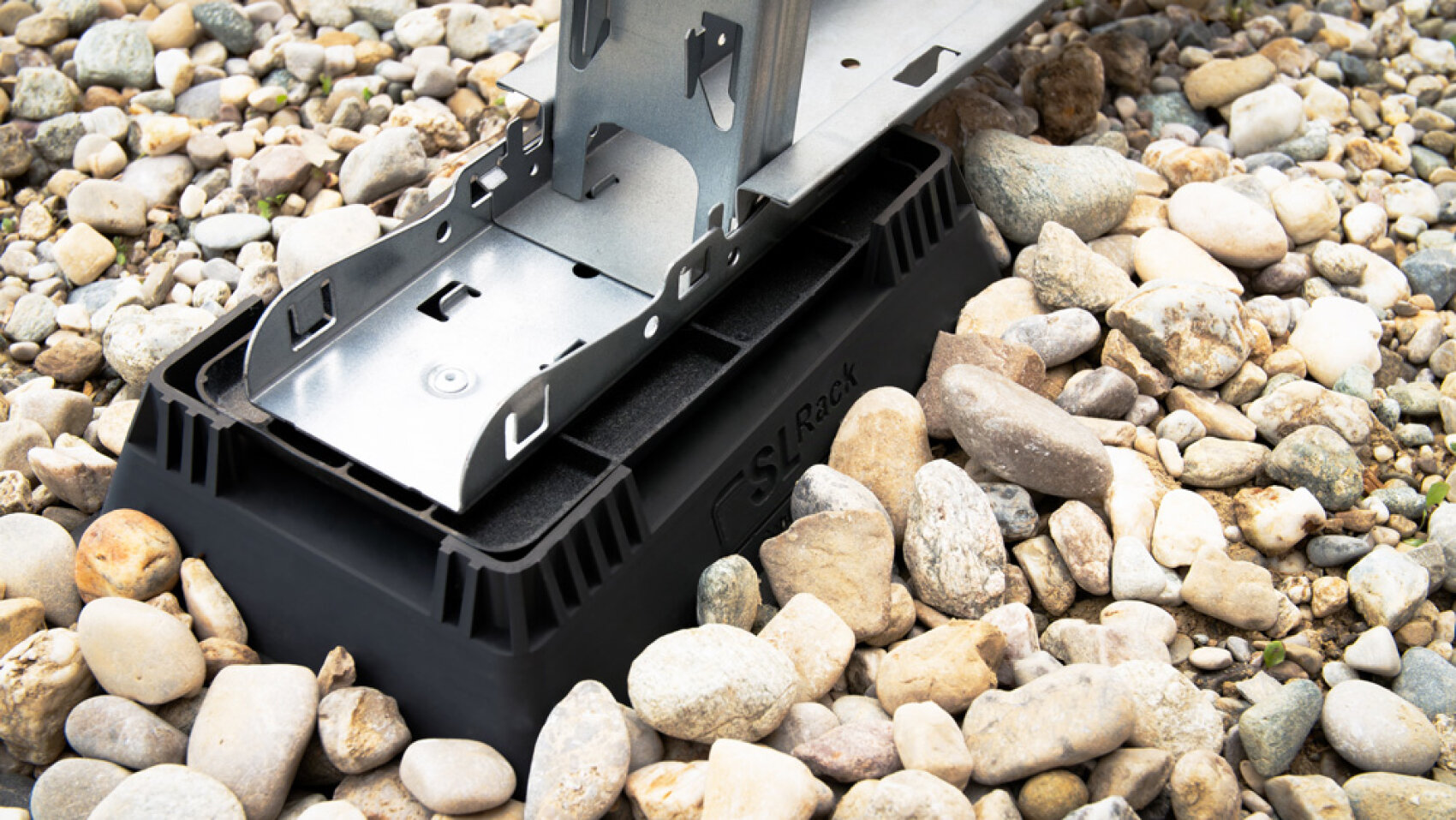 Nahaufnahme einer Metallhalterung auf einem schwarzen Kunststoffsockel, die auf einem Bett aus kleinen, hellen Steinen im Freien steht.